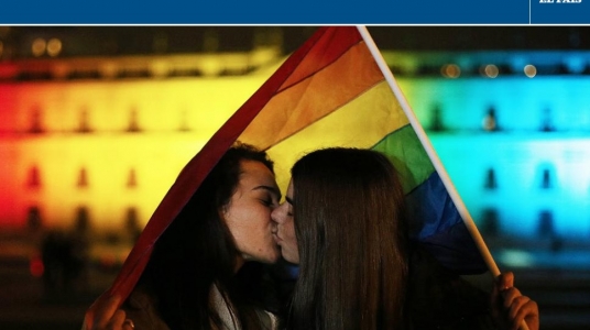 Pareja besándose con bandera del orgullo LGBTQI detrás. Imagen de El País.